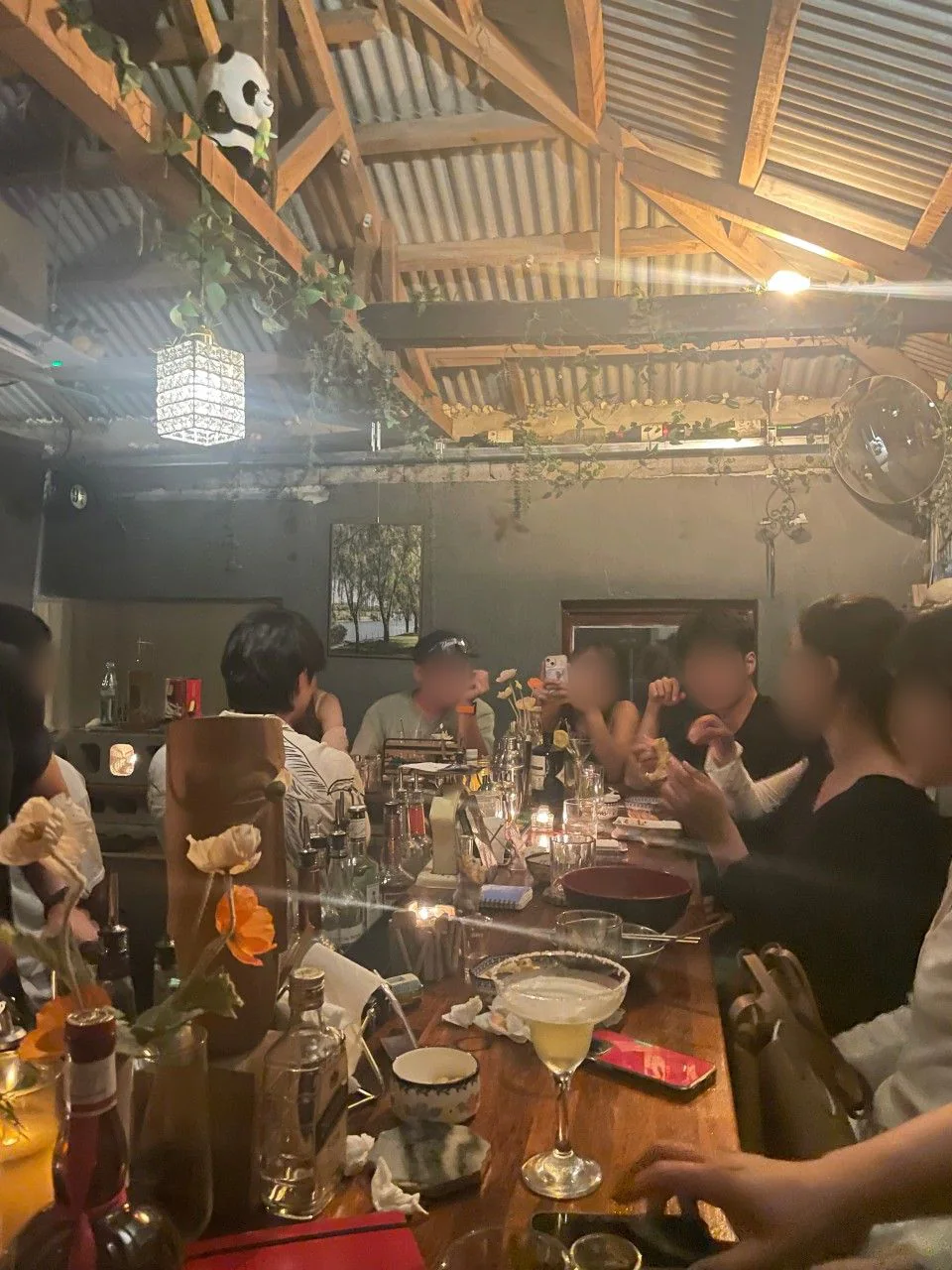 Guests seated around the long wooden bar counter at Jeju Ahop Solo Bar Sillim, under exposed rafters, a corrugated roof and a crystal lamp